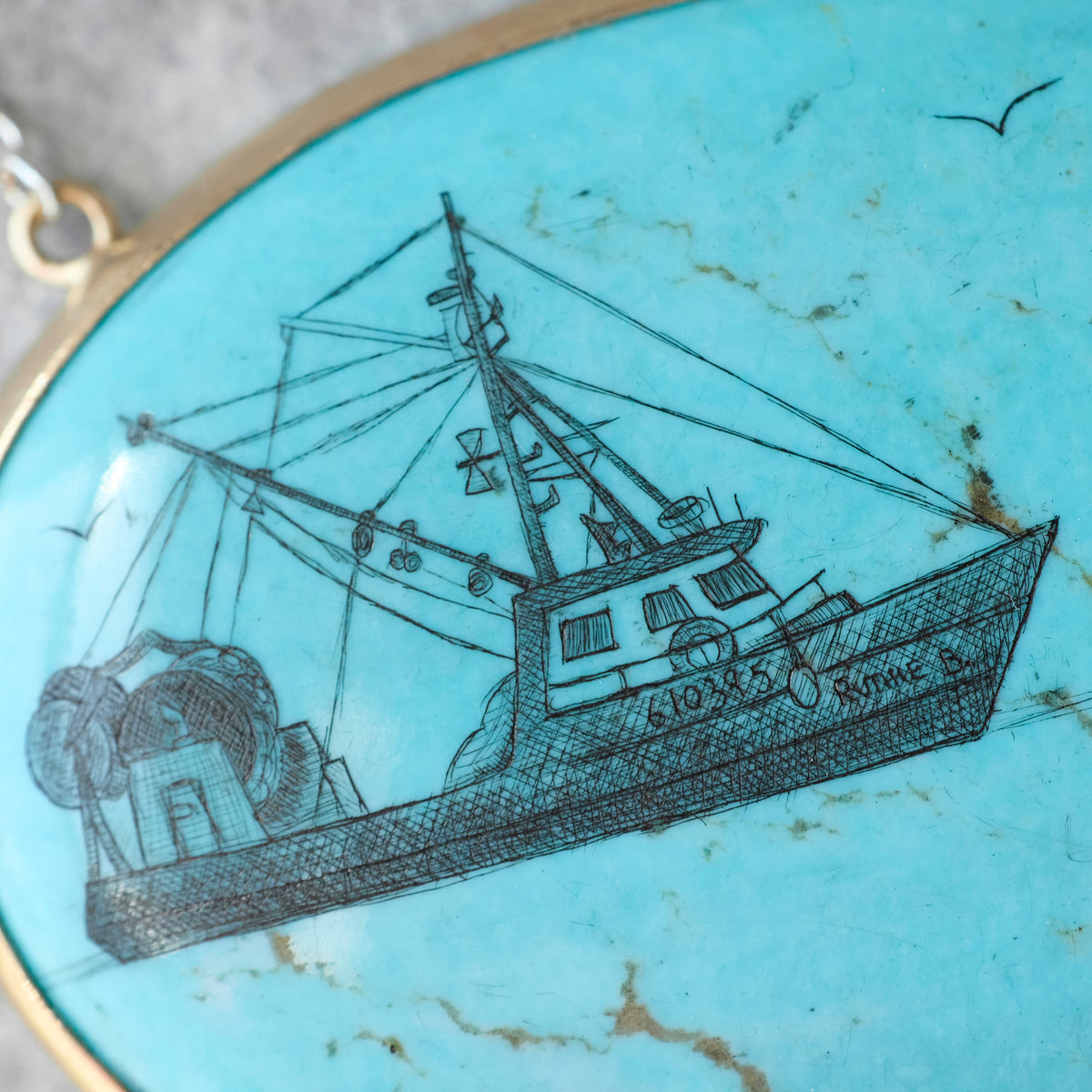 A close-up of a round turquoise pendant featuring a detailed black ink sketch of a fishing boat named 'SIOX X KINGS B'.