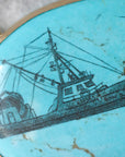 A close-up of a round turquoise pendant featuring a detailed black ink sketch of a fishing boat named 'SIOX X KINGS B'.