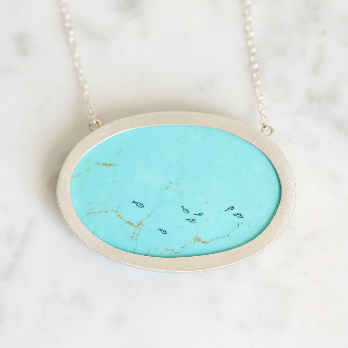 An oval pendant necklace featuring a vibrant turquoise stone inlay with natural gold and dark fissures, set in a brushed silver-tone metal frame and suspended from a delicate silver chain, resting on a white marble surface.