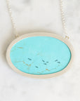 An oval pendant necklace featuring a vibrant turquoise stone inlay with natural gold and dark fissures, set in a brushed silver-tone metal frame and suspended from a delicate silver chain, resting on a white marble surface.