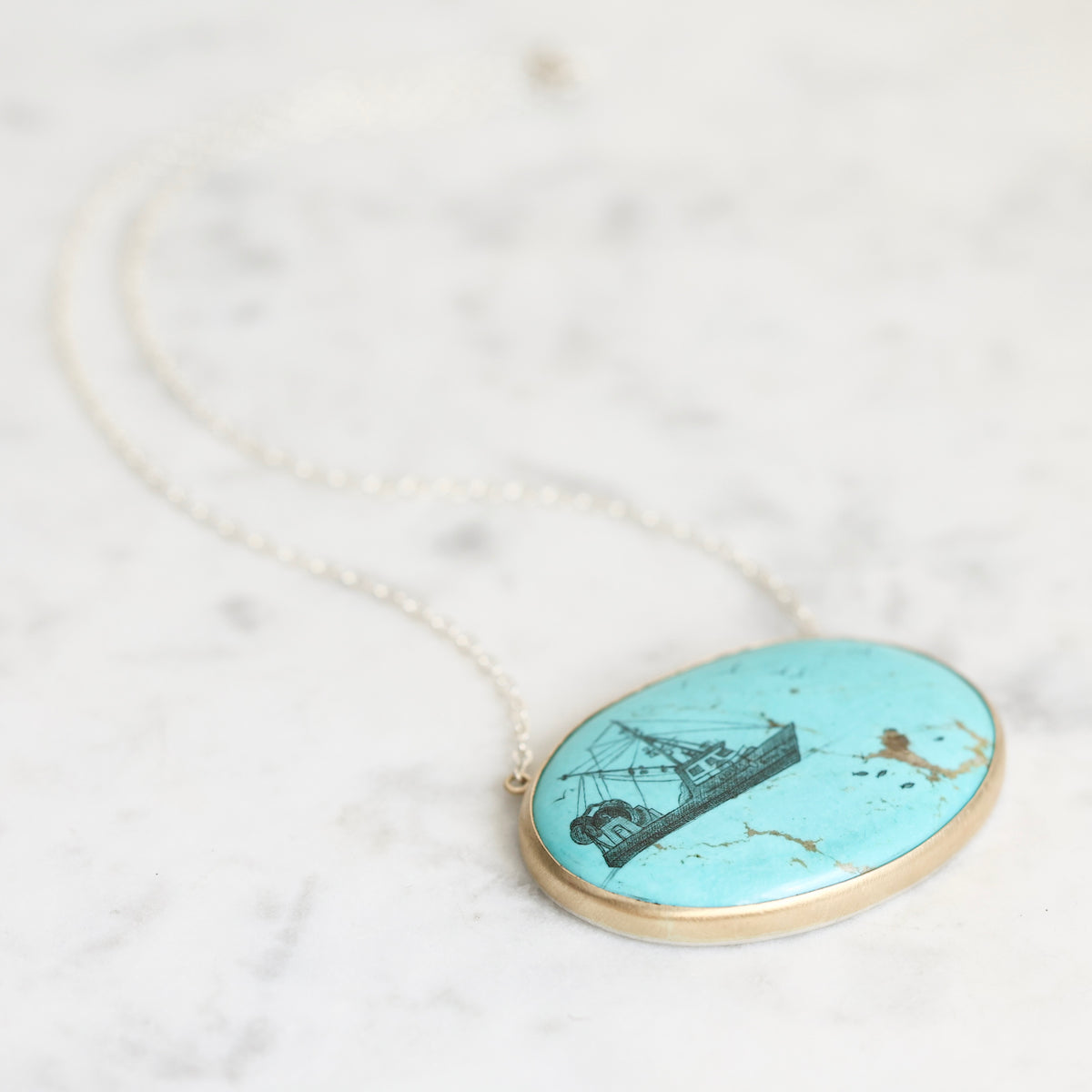 An oval pendant necklace featuring a turquoise-colored stone with a black engraving of a sailing ship, set in a gold-tone bezel on a delicate silver-tone chain, displayed on a light marble surface.