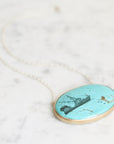 An oval pendant necklace featuring a turquoise-colored stone with a black engraving of a sailing ship, set in a gold-tone bezel on a delicate silver-tone chain, displayed on a light marble surface.