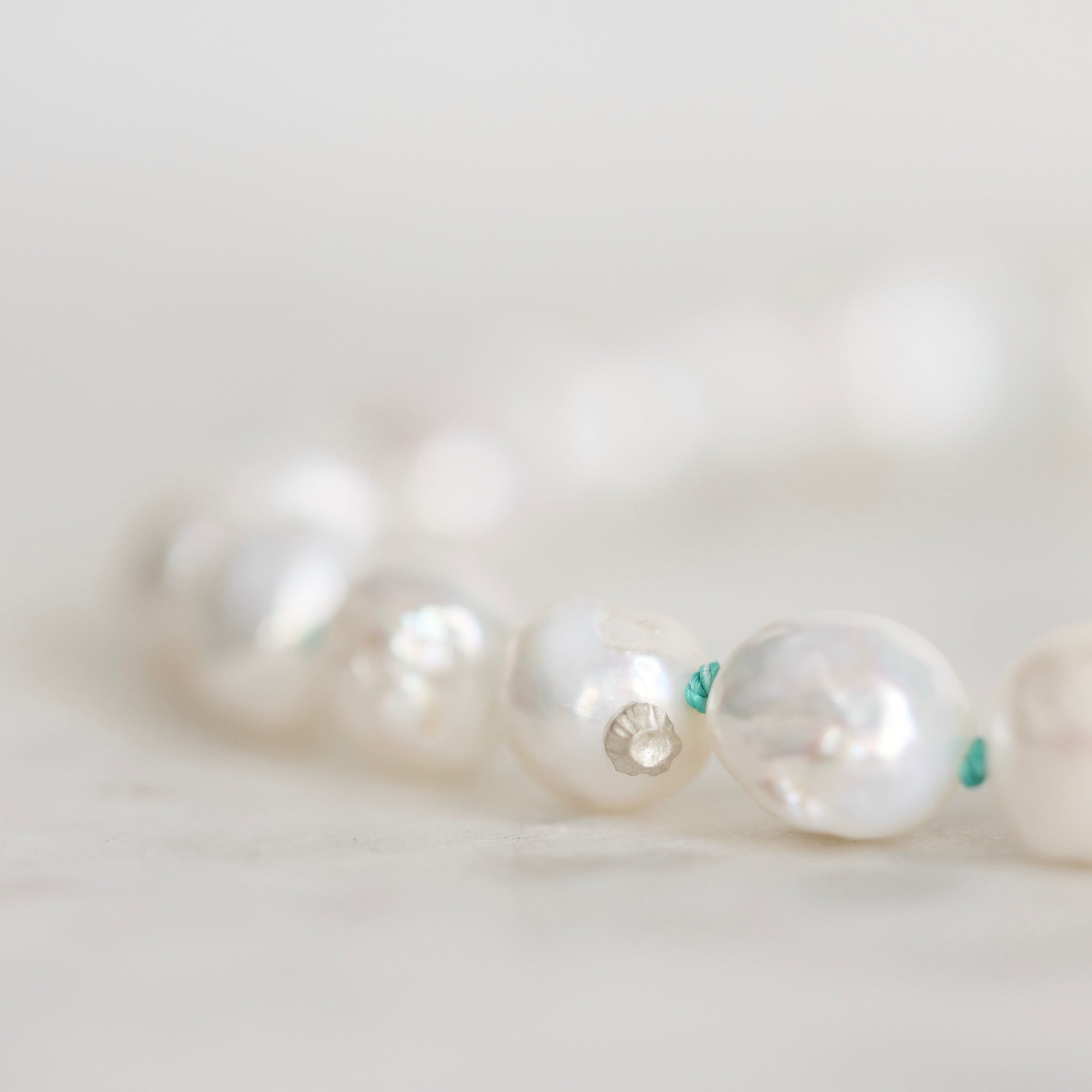 A close-up of a beaded strand featuring irregular, lustrous white pearls separated by tiny green string, photographed on a soft light background.