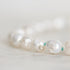 A close-up of a beaded strand featuring irregular, lustrous white pearls separated by tiny green string, photographed on a soft light background.
