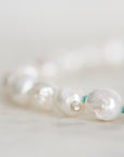 A close-up of a beaded strand featuring irregular, lustrous white pearls separated by tiny green string, photographed on a soft light background.