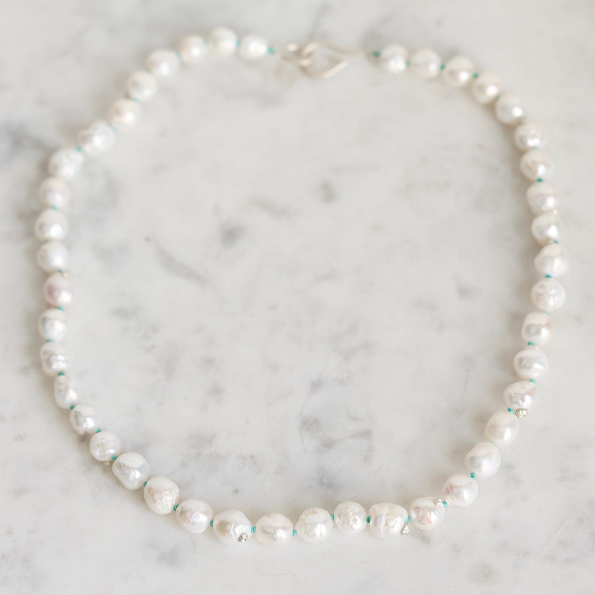 A strand of irregular white freshwater pearls with small turquoise blue spacer beads, arranged in a circle on a light gray marble surface.