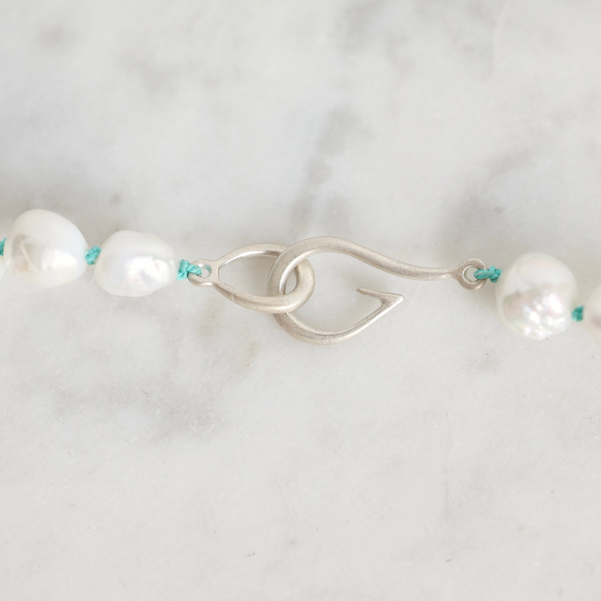 A close-up of a pearl necklace with a silver fishhook clasp and turquoise cord accents, laid on a light marble surface.