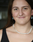 A woman with brown hair wearing a white baroque pearl necklace with a silver clasp and matching blue teardrop earrings, shown in a close-up portrait.