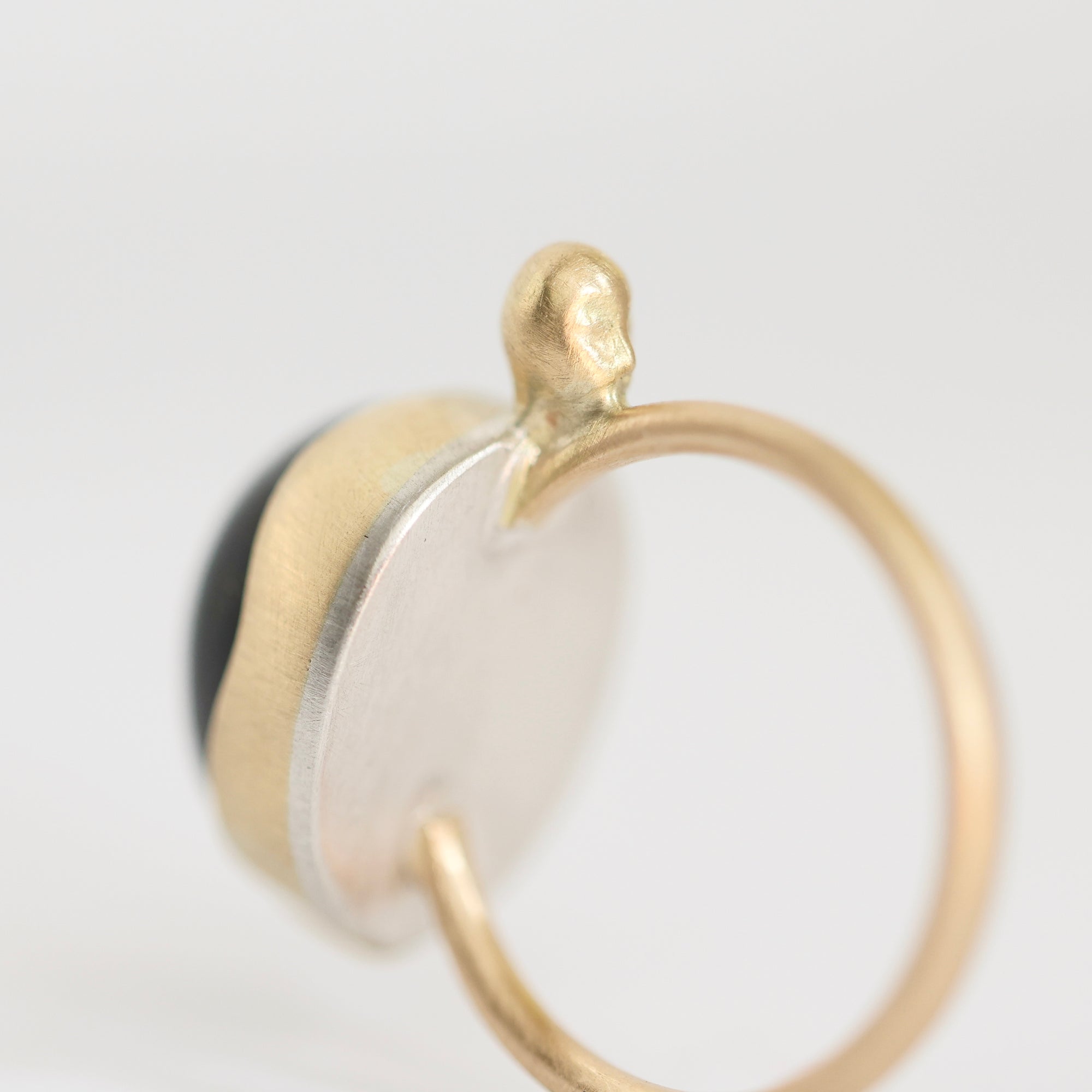 A close-up of a gold ring with a textured circular gold and silver-toned face, featuring a small gold protruding bead on top and a dark inlay on the side, set against a plain white background.