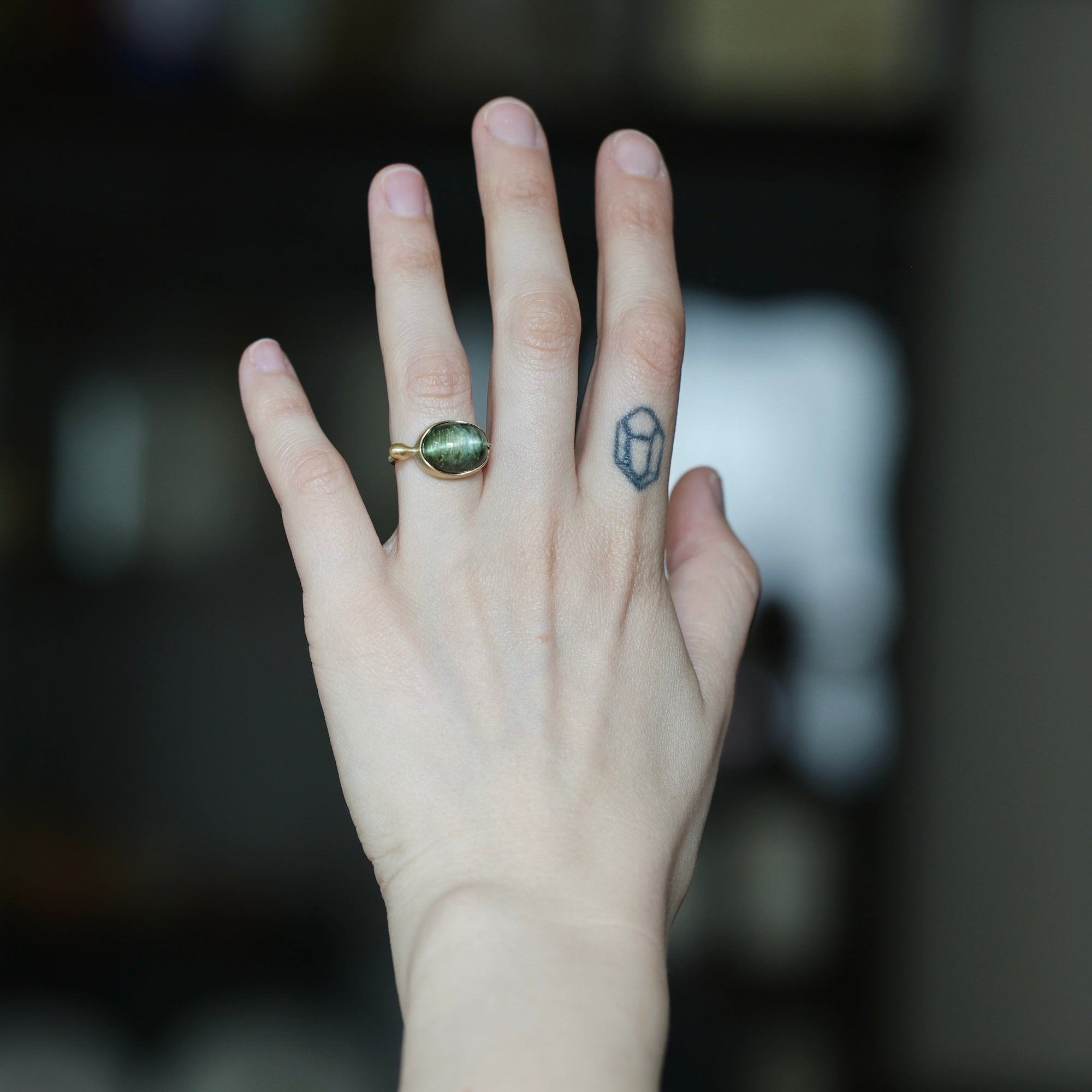 A hand wearing a gold ring with an oval green stone, shown against a dark, blurred background. The hand also features a small geometric tattoo on one of the fingers.