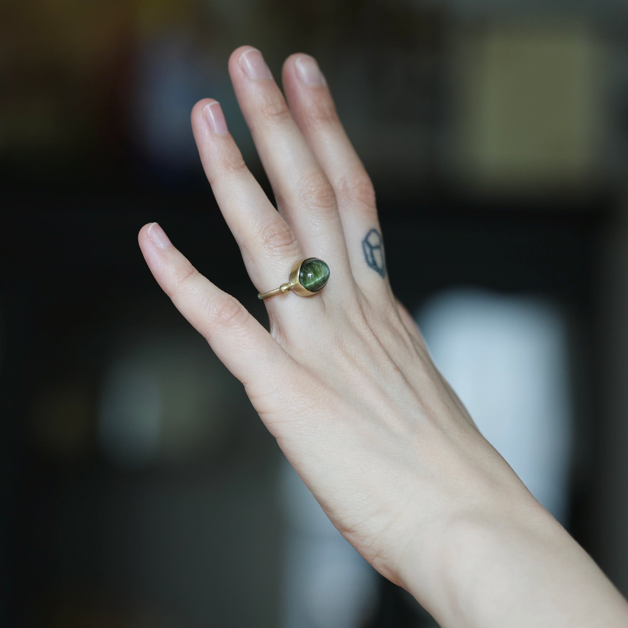 A gold ring with a round green gemstone worn on a person's finger, shown against a dark, blurred background.