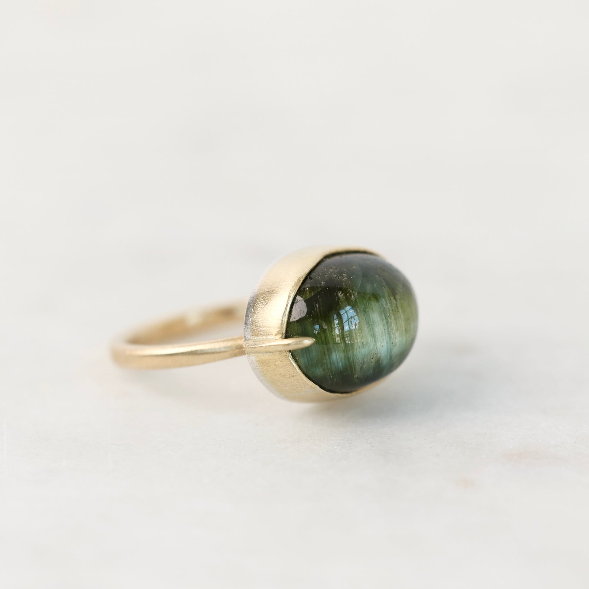 A gold ring with a bezel-set oval green gemstone, photographed on a light gray background.