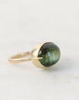 A gold ring with a bezel-set oval green gemstone, photographed on a light gray background.