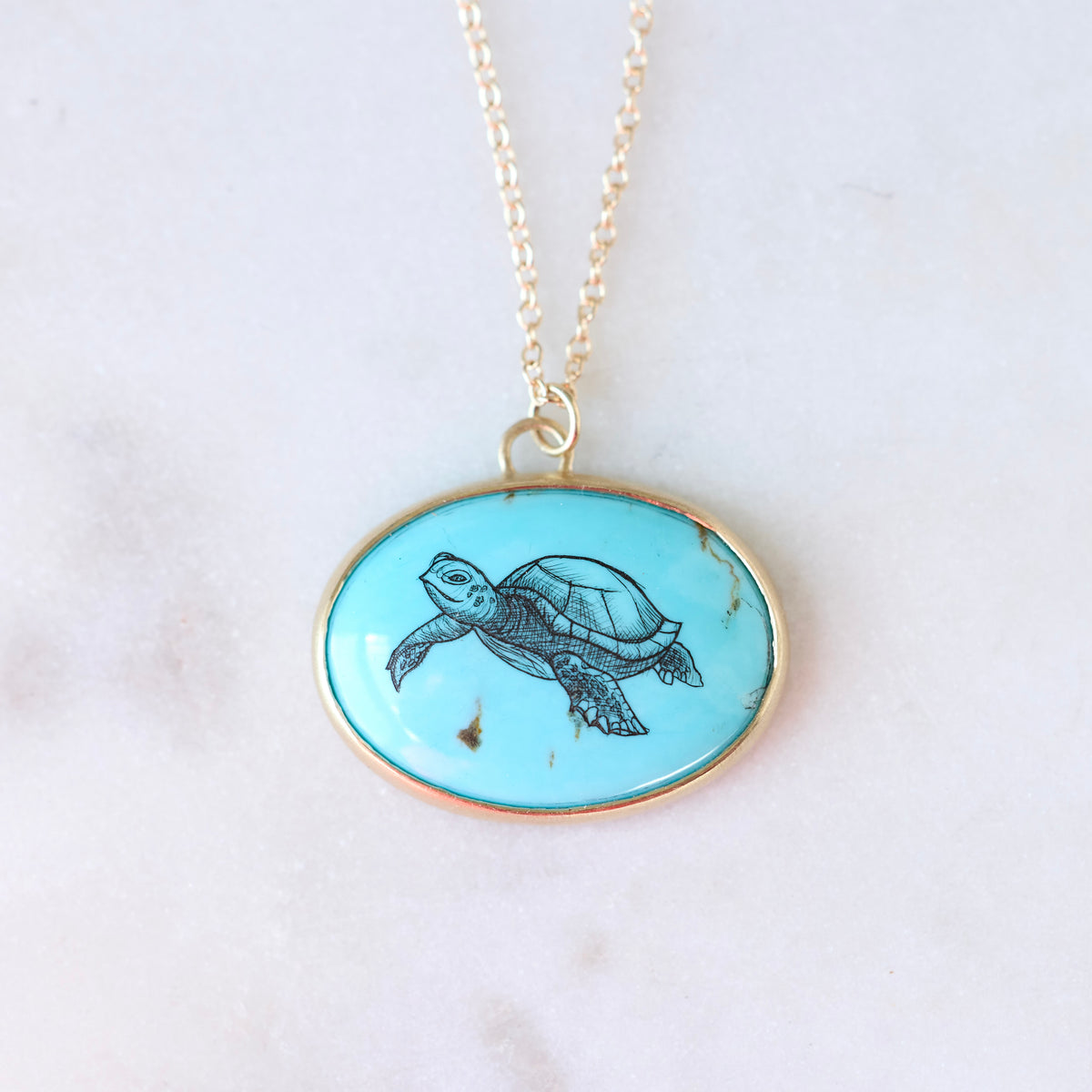 An oval pendant necklace featuring a turquoise blue background with a black ink-style illustration of a sea turtle, set in a gold-tone bezel and hanging from a gold chain on a light marble surface.