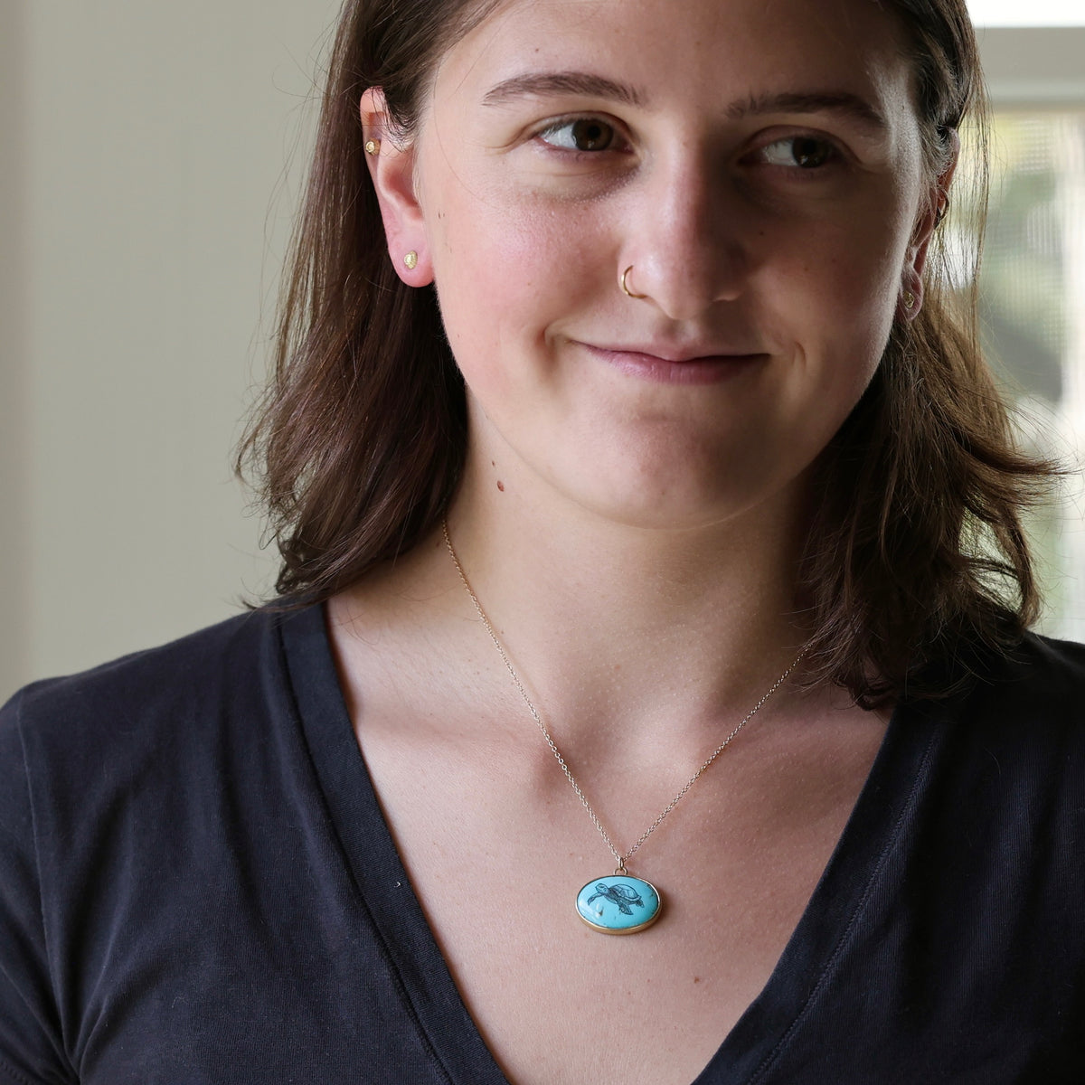 A woman wearing a delicate gold chain necklace with a round turquoise pendant featuring a classical-style portrait.