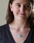 A woman wearing a delicate gold chain necklace with a round turquoise pendant featuring a classical-style portrait.