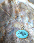 A gold chain necklace with an oval pendant featuring a black turtle illustration on a light turquoise background, displayed on a textured abalone shell.