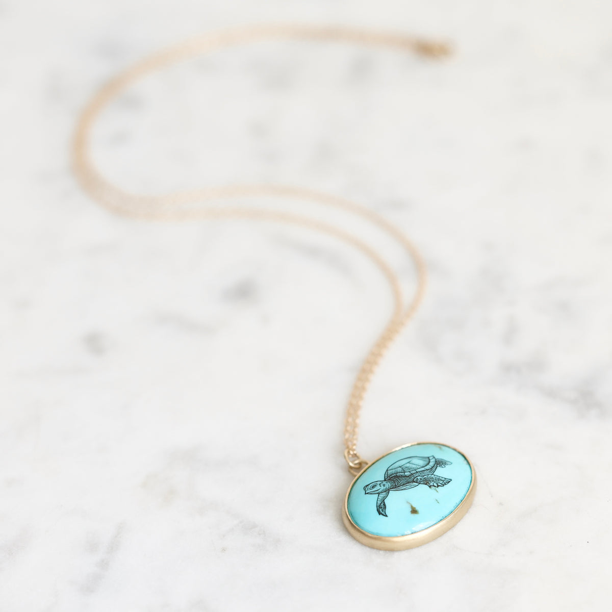 A gold chain necklace with an oval pendant featuring a turquoise background and a black turtle illustration, resting on a light marble surface.