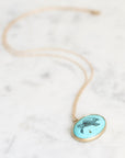 A gold chain necklace with an oval pendant featuring a turquoise background and a black turtle illustration, resting on a light marble surface.