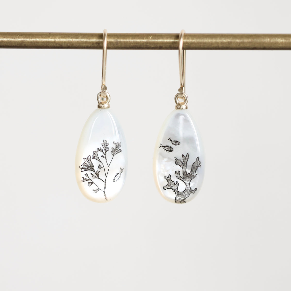 A pair of gold-tone drop earrings with teardrop-shaped white mother-of-pearl pendants featuring black botanical and fish illustrations, hanging from a gold rod against a light gray background.