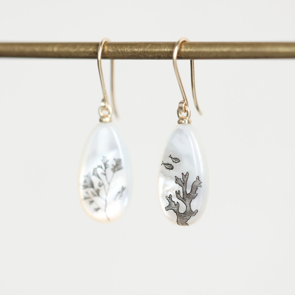 A pair of teardrop-shaped white earrings with gold fishhook wires, hanging from a gold rod. One earring contains dried botanical elements, and the other features a small fish and coral design.