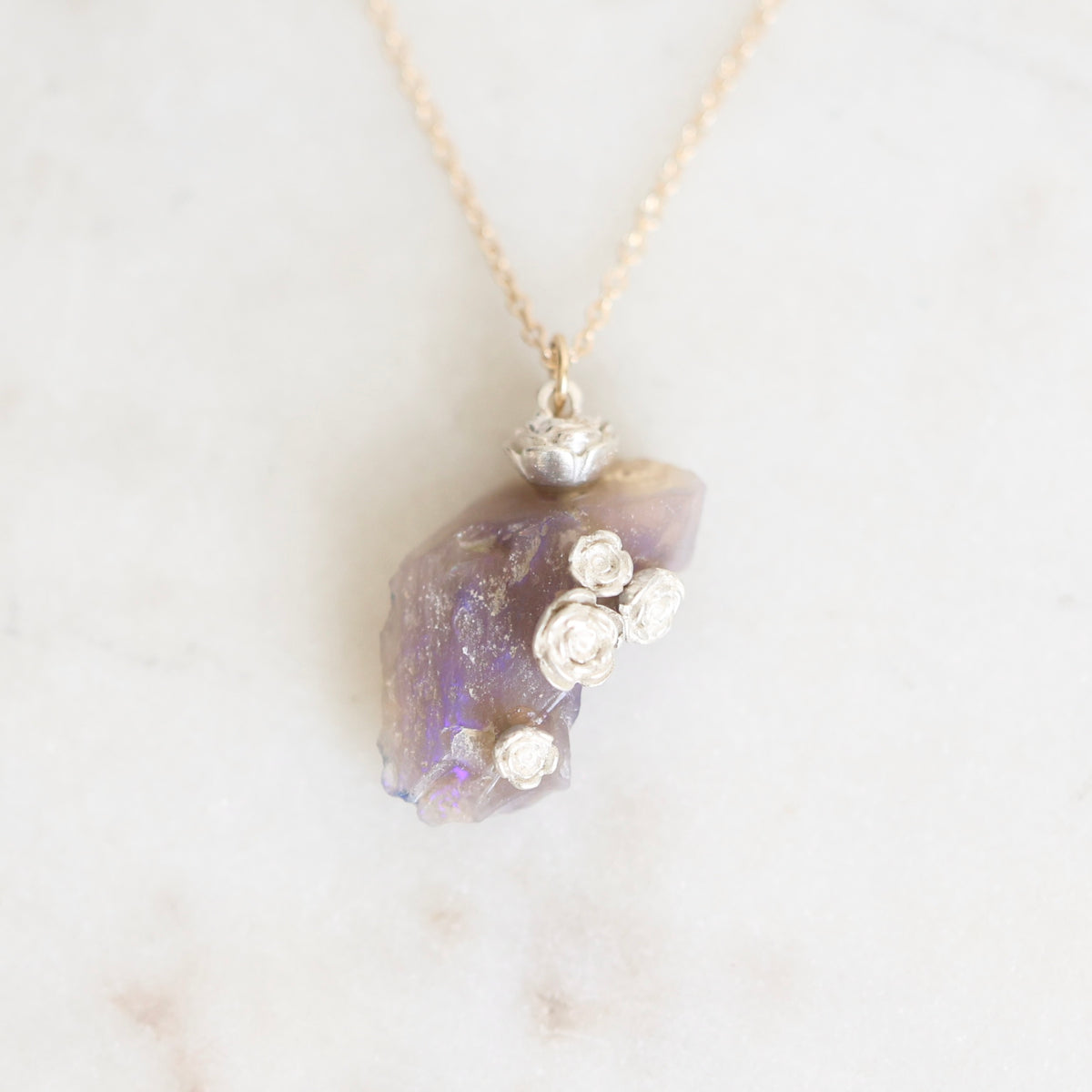 Bright purple fossilized opal with silver rose details suspended from a gold chain against a marble background. 
