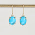 A pair of oval turquoise drop earrings with gold prong settings and gold hooks, hanging from a gold display bar against a light gray background.