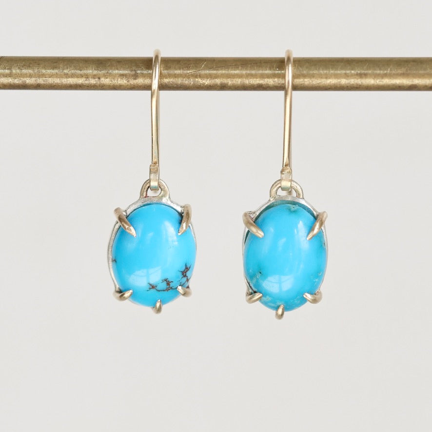 A pair of oval turquoise drop earrings with gold prong settings and gold hooks, hanging from a gold display bar against a light gray background.