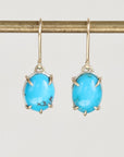 A pair of oval turquoise drop earrings with gold prong settings and gold hooks, hanging from a gold display bar against a light gray background.