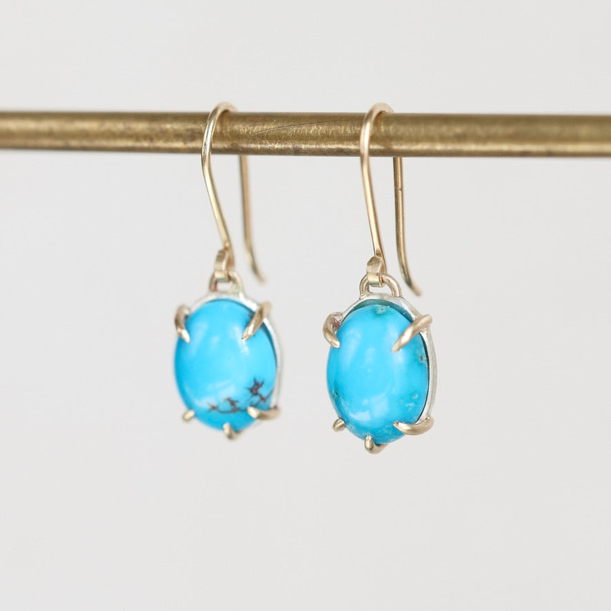 A pair of gold drop earrings with oval turquoise-blue gemstones in a prong setting, hanging from a gold rod against a light gray background.