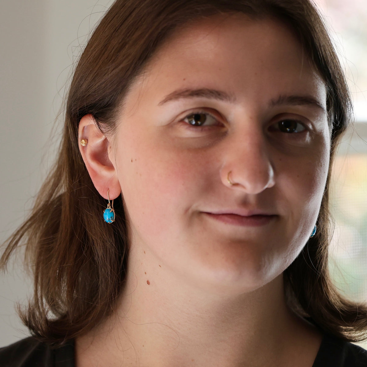 A close-up portrait of a woman with brown hair wearing small gold hoop earrings with bright blue teardrop-shaped stones.