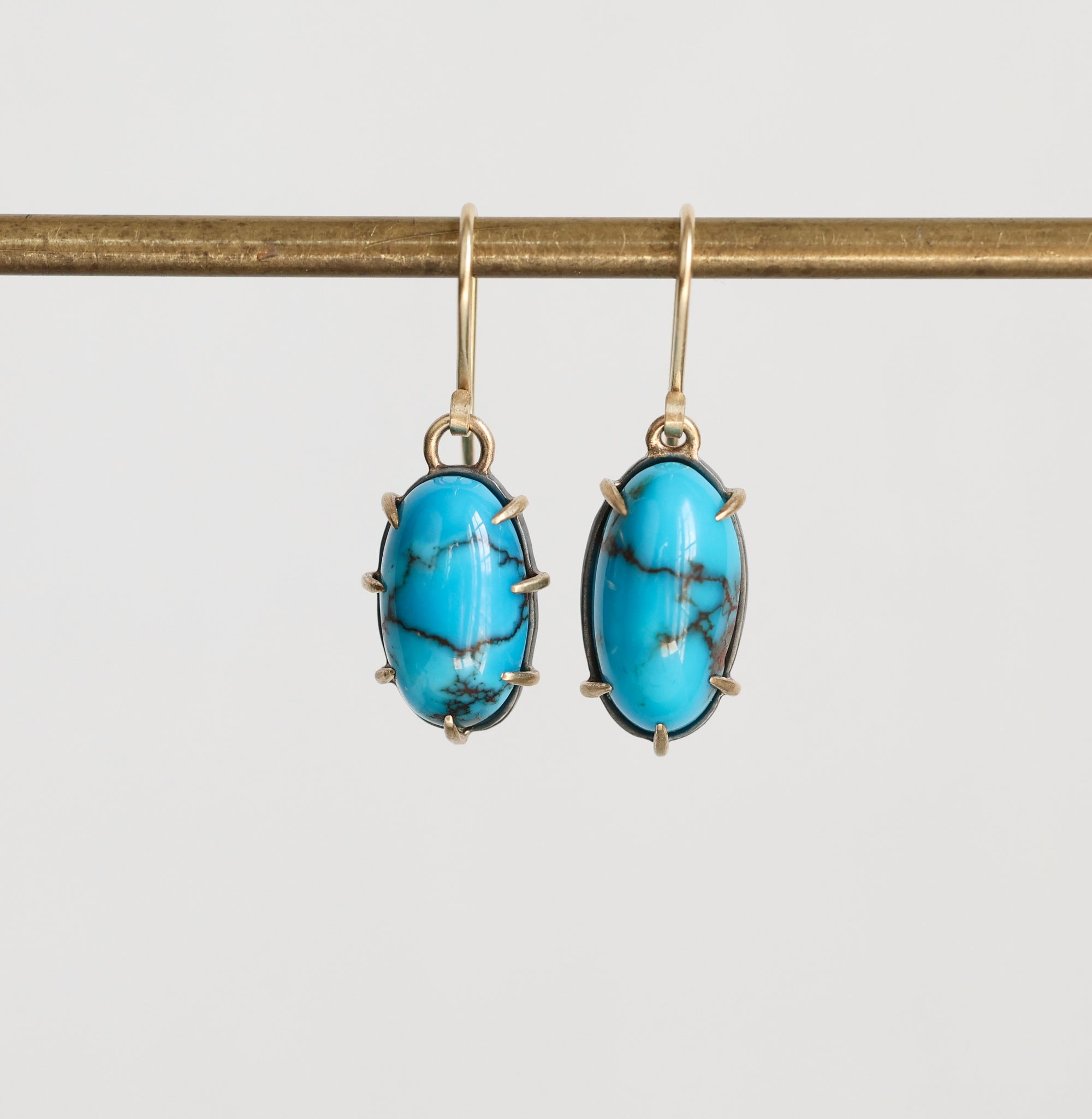 A pair of oval drop earrings with bright turquoise blue marbled stones in gold-toned bezel settings, hanging from gold hooks against a light gray background.