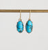 A pair of oval drop earrings with bright turquoise blue marbled stones in gold-toned bezel settings, hanging from gold hooks against a light gray background.