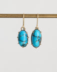 A pair of oval drop earrings with bright turquoise blue marbled stones in gold-toned bezel settings, hanging from gold hooks against a light gray background.