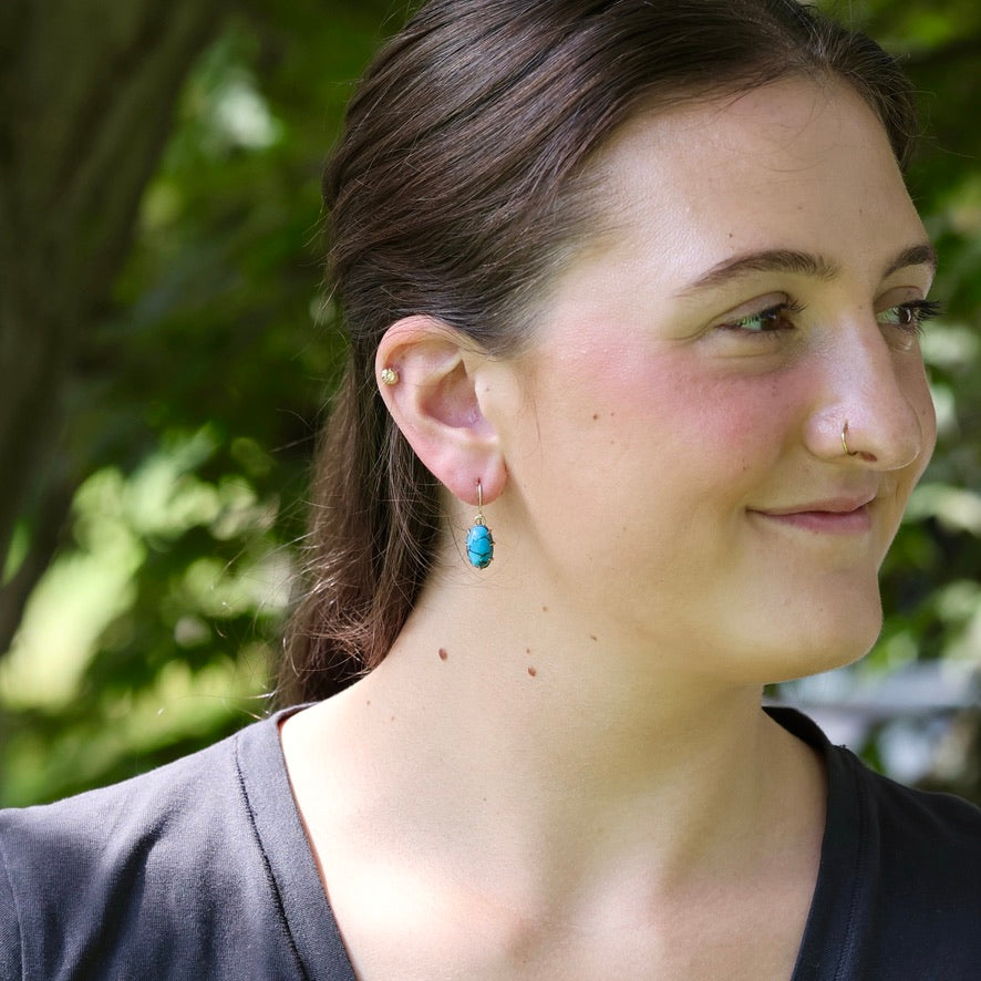 A close-up side profile of a woman outdoors wearing a small turquoise drop earring with a gold hoop.