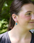 A close-up side profile of a woman outdoors wearing a small turquoise drop earring with a gold hoop.