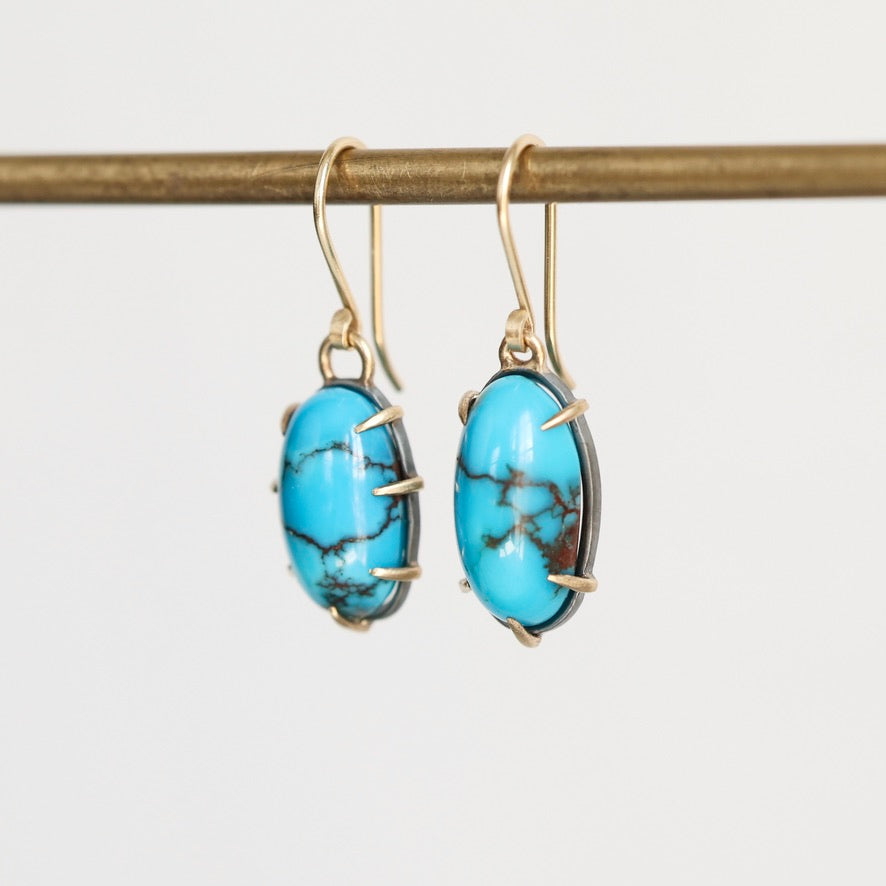 A pair of gold drop earrings with oval turquoise-blue marbled stones, hanging from a gold rod against a light gray background.