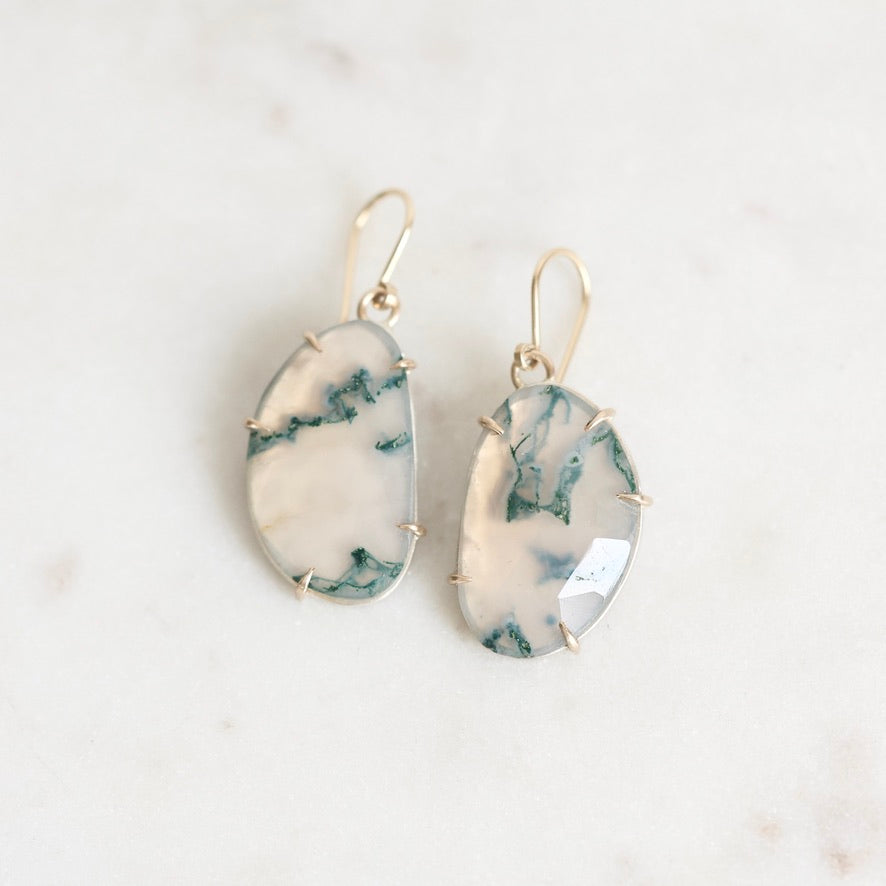 A pair of gold drop earrings with irregular organic-shaped cabochon gemstones featuring a white and dark green marbled pattern, displayed on a light marble surface.