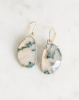 A pair of gold drop earrings with irregular organic-shaped cabochon gemstones featuring a white and dark green marbled pattern, displayed on a light marble surface.