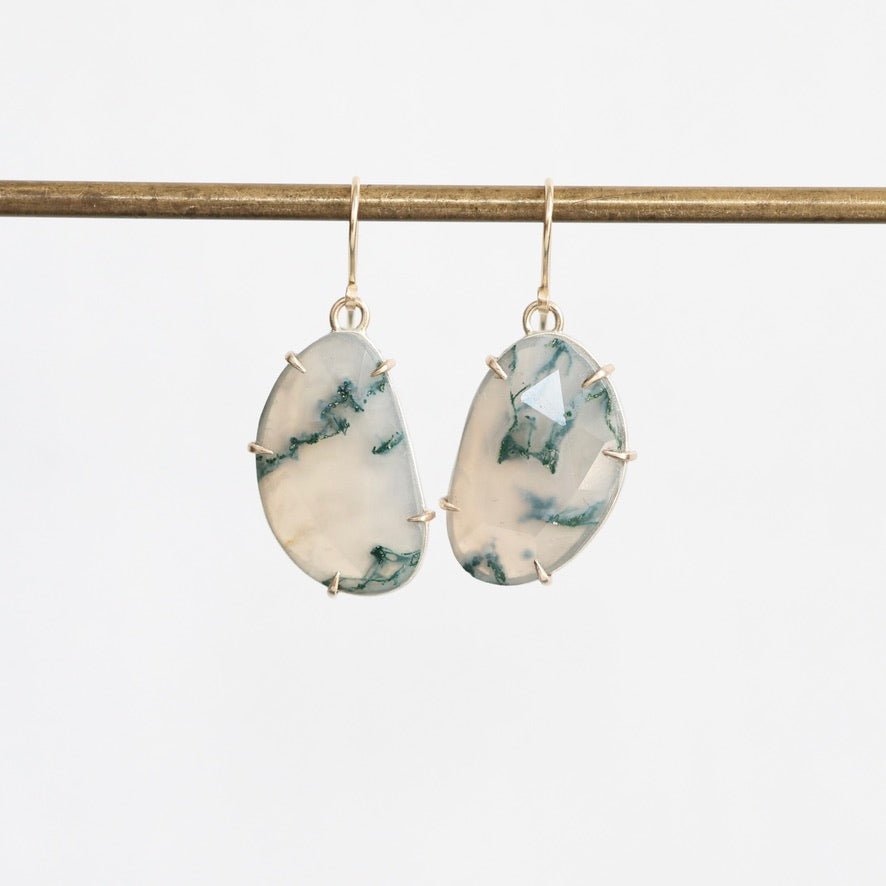 A pair of teardrop-shaped drop earrings with white and green marbled stone inlays set in gold-toned metal, hanging from a gold-toned horizontal bar against a light gray background.