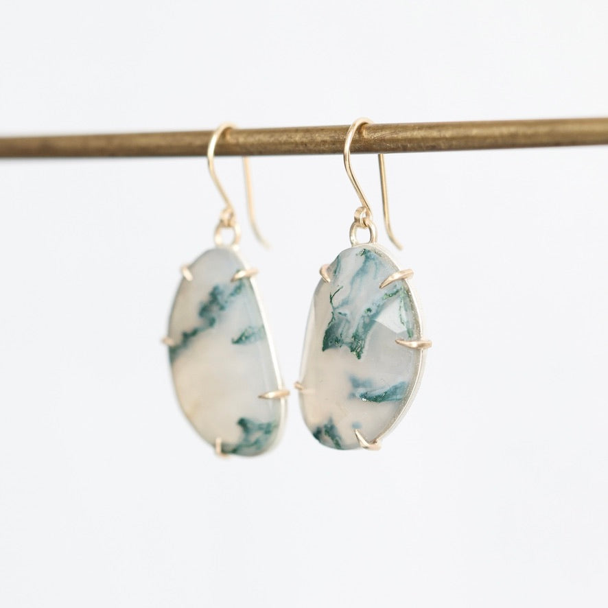 A pair of gold drop earrings with irregular white and green marbled stone pendants, hanging from a gold-colored horizontal bar against a plain white background.