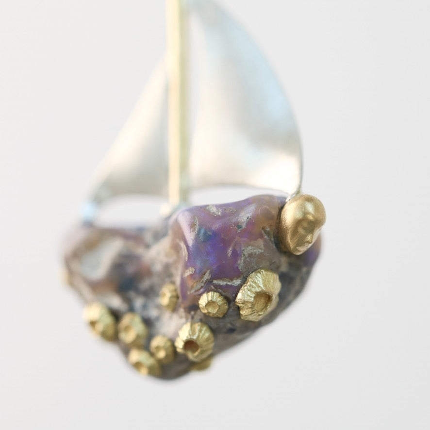 A close-up of a decorative sailboat ornament or figurine with a metallic silver hull and sail, a purple iridescent center, and raised gold accents on a plain light background.