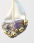 A close-up of a decorative sailboat ornament or figurine with a metallic silver hull and sail, a purple iridescent center, and raised gold accents on a plain light background.