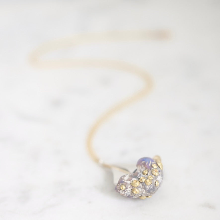 A delicate gold chain necklace with a small, organic-shaped pendant covered in tiny gold rhinestones and a subtle iridescent lavender-blue center, resting on a white marble surface.