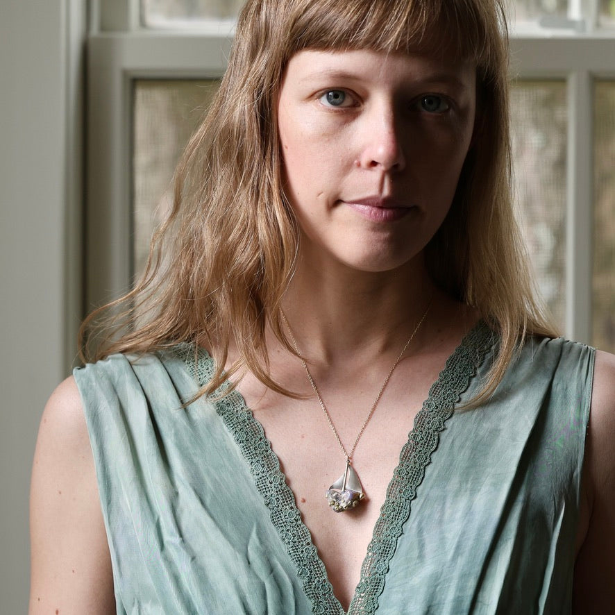 A woman with light brown hair and blue eyes wearing a sage green lace-trimmed top and a silver pendant necklace, photographed in a softly lit indoor setting.