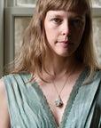 A woman with light brown hair and blue eyes wearing a sage green lace-trimmed top and a silver pendant necklace, photographed in a softly lit indoor setting.