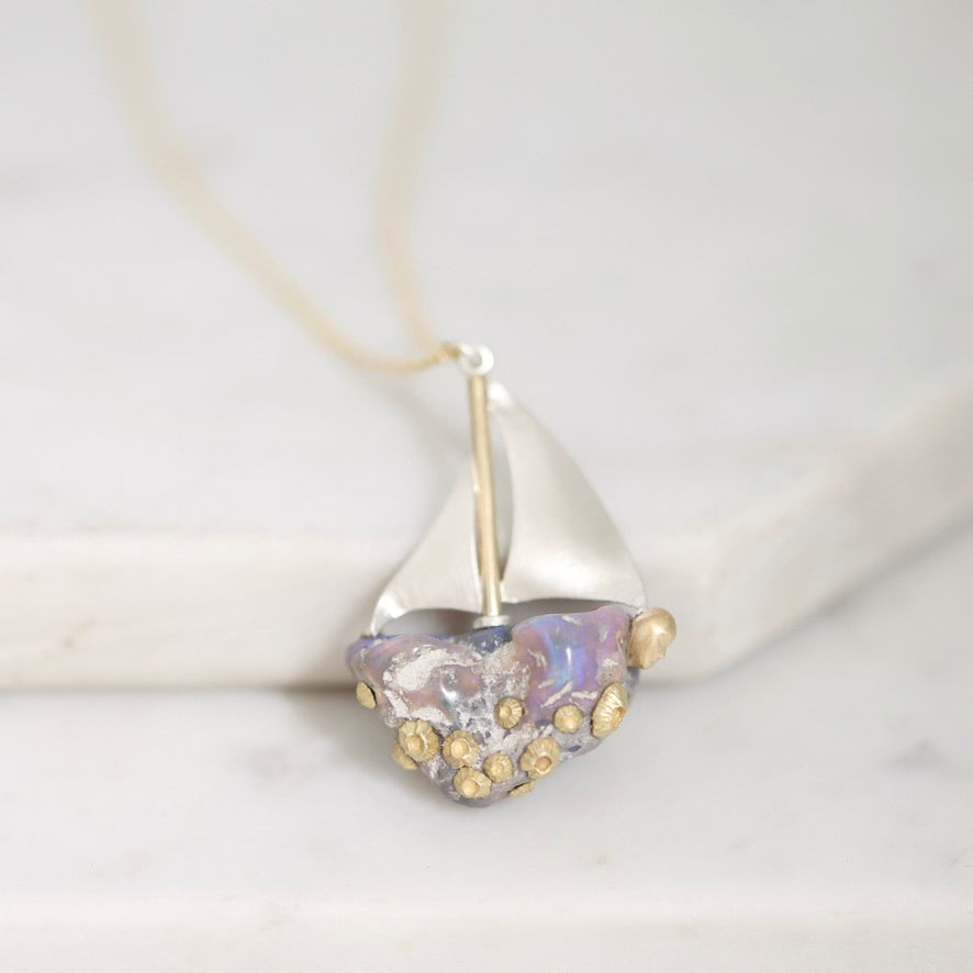 A close-up of a delicate necklace featuring a sailboat-shaped pendant with a silver hull, gold accents, and a translucent iridescent heart-shaped glass center, resting on a light marble surface.