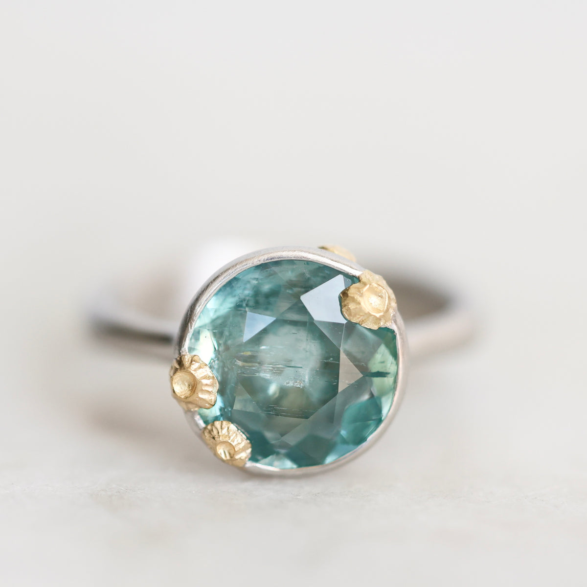 A close-up shot of a ring featuring a round faceted light blue-green gemstone in a silver-toned setting with three small gold-toned accents, resting on a light neutral surface.