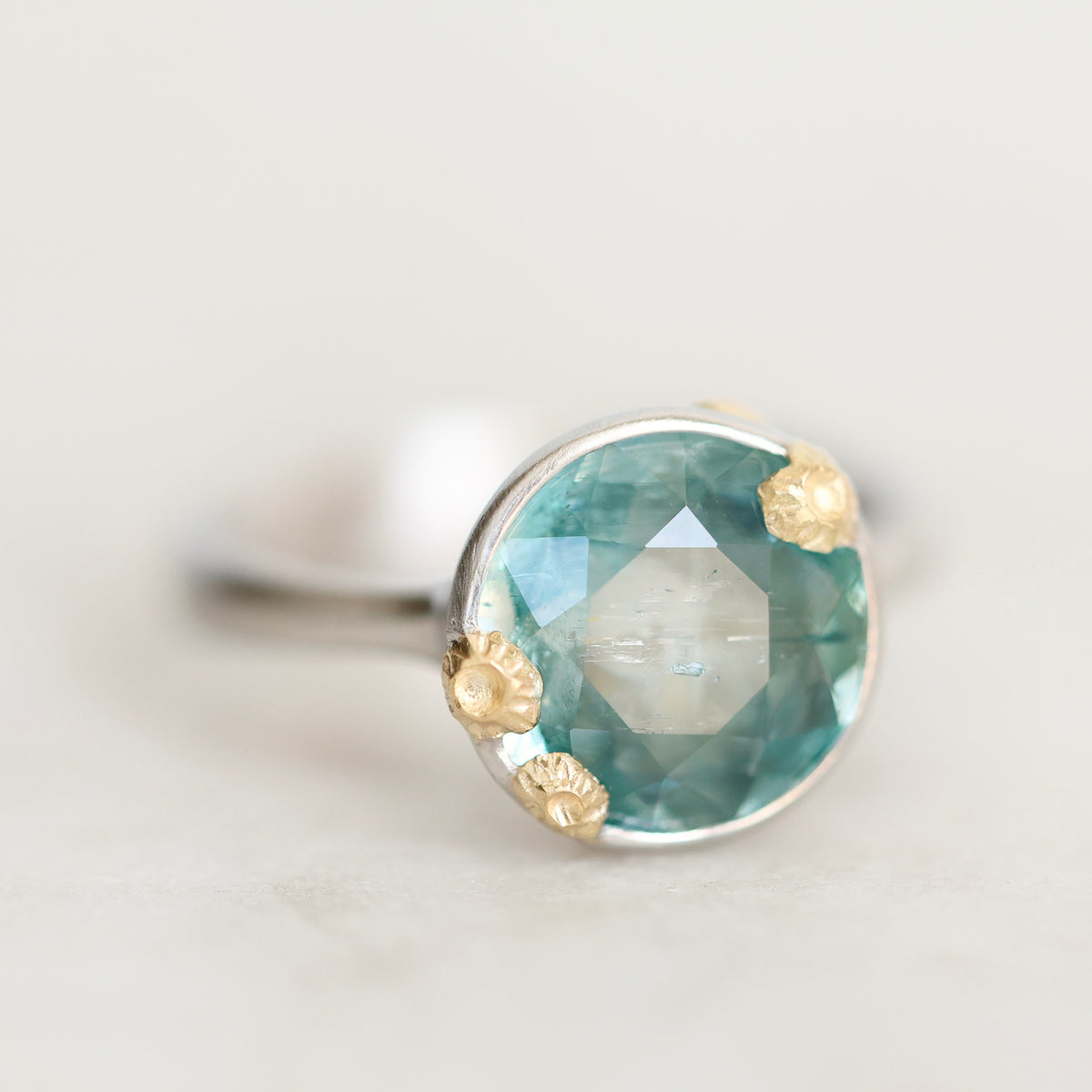 A close-up shot of a ring featuring a round faceted aquamarine gemstone in a bezel setting with textured gold accents on a silver band, set against a soft, light gray background.