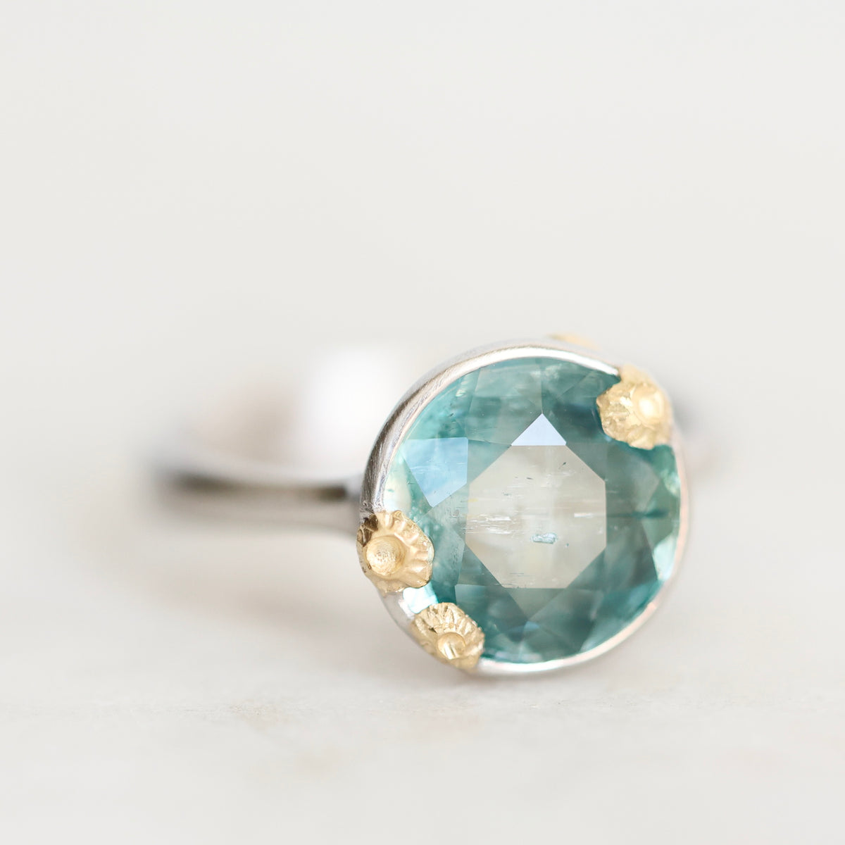 A close-up shot of a ring featuring a faceted light blue-green gemstone in a silver-toned setting with three small gold-toned flower-like accents, on a light neutral background.